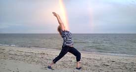 Diane Kovanda doing yoga on a beach with a rainbow in the background.